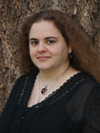 white woman with fluffy brown wavy hair, wearing a black shirt, behind her the tunk of a large fir tree