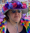 me at pride, white woman in sunglasses with a rainbow shirt on sholders and wearing a hat with large magenta, blue and purple flowers